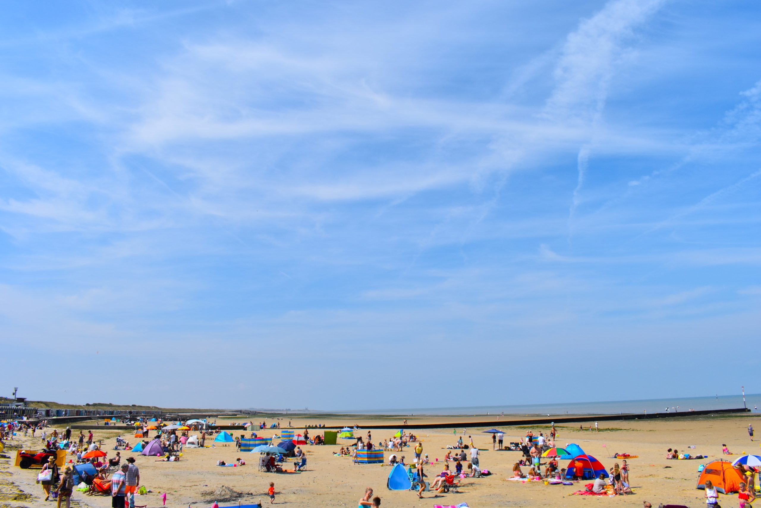 Flat sandy beach with calm sea, promenade, and a few people enjoying a sunny day
