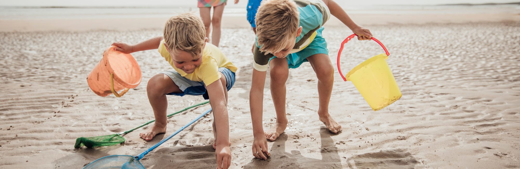 Two young boys with colourful buckets exploring wet sand and shallow pools at low tide on a sunny beach