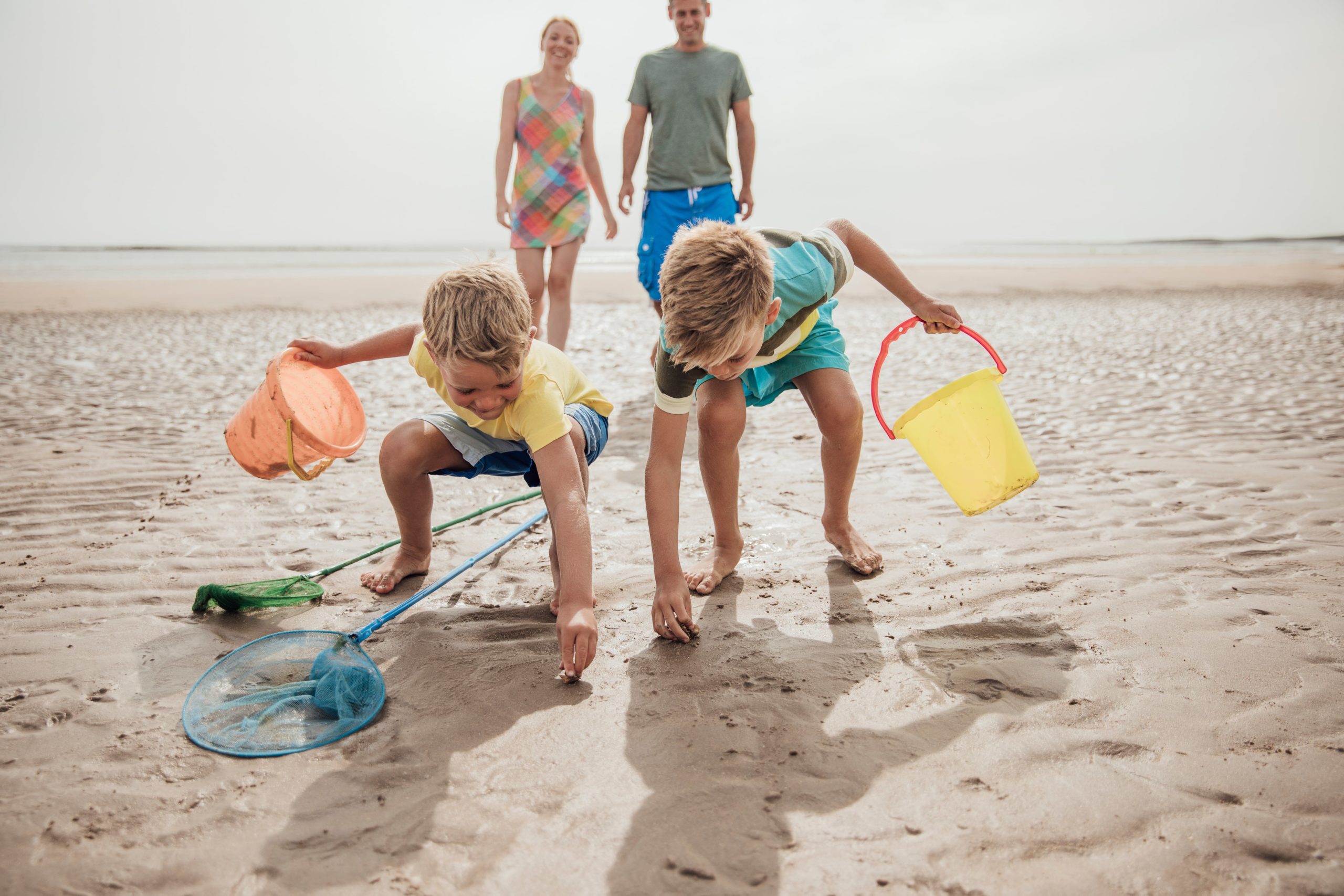 Two young boys with colourful buckets exploring wet sand and shallow pools at low tide on a sunny beach