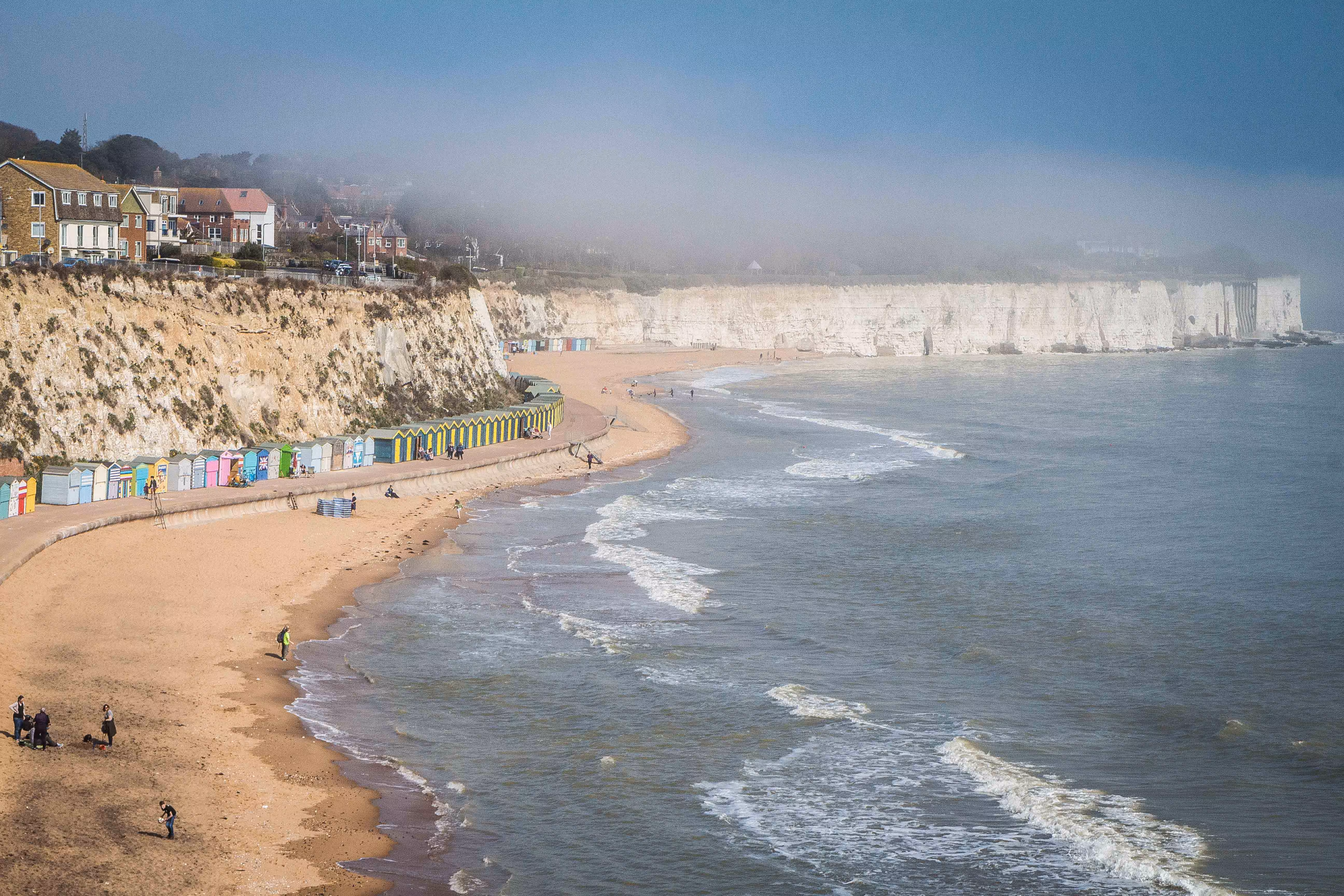 A misty coastal beach in Broadstairs with golden sand, colourful beach huts, and dramatic white chalk cliffs.